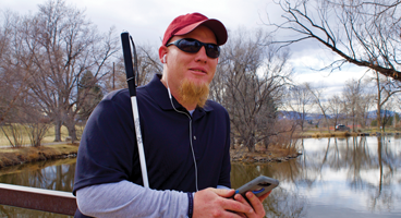 A young blind man wearing ear buds, listens to NFB-NEWSLINE on his phone outside.